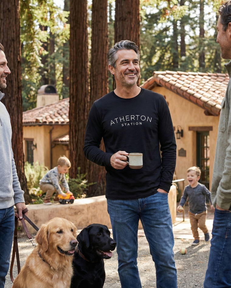 Man in black 'Atherton Station' t-shirt holding a mug with two dogs and children in the background.