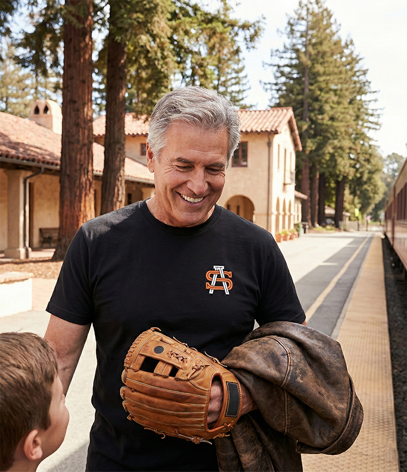 Man holding a baseball glove and jacket on a train platform with trees and a building in the background