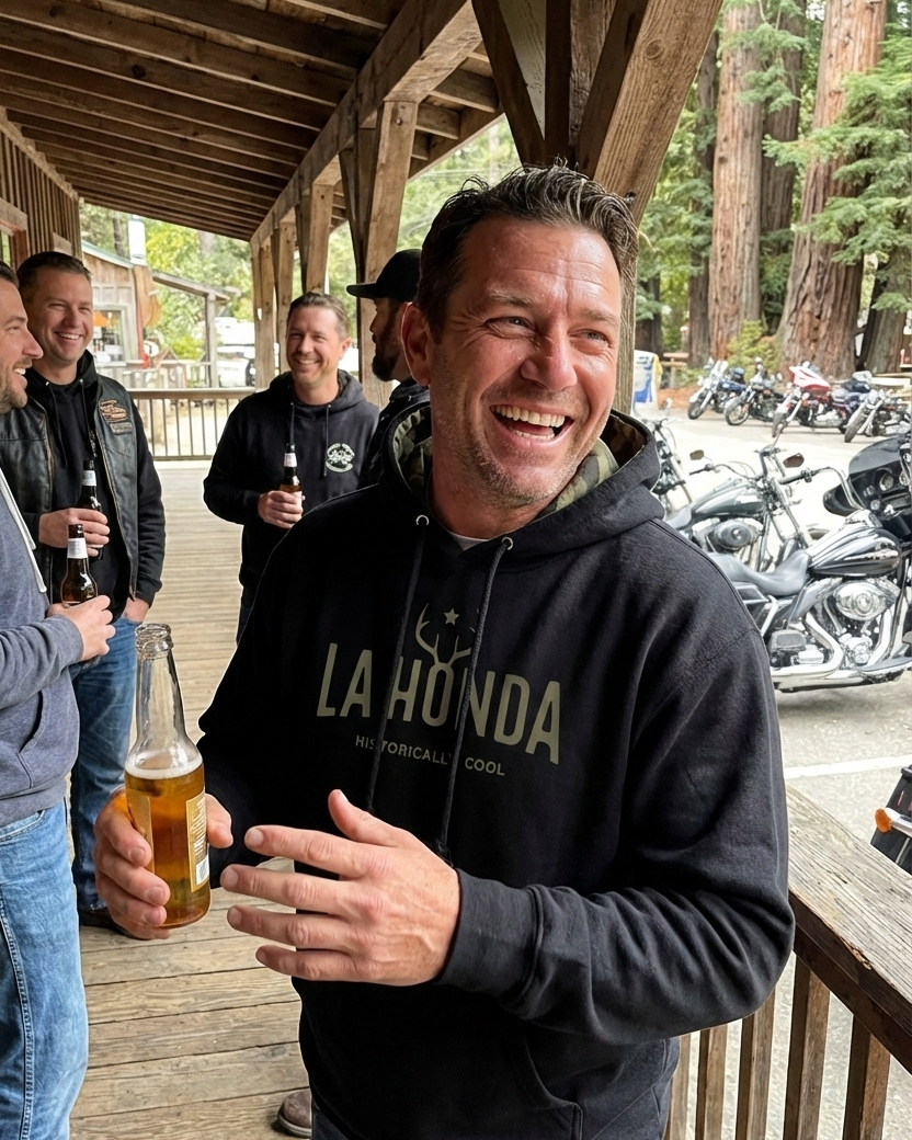 Man in a 'La Honda' hoodie holding a beer on a wooden deck with motorcycles and people in the background.