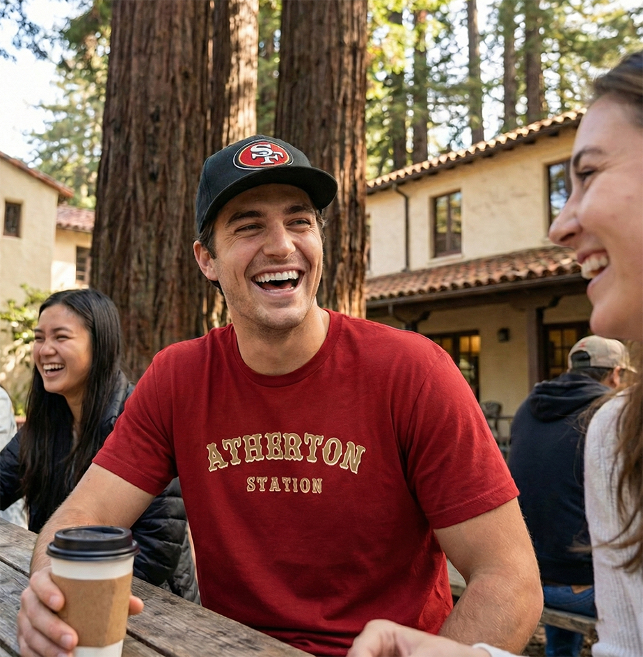 Man in red 'Atherton Station' t-shirt and SF 49ers cap laughing with friends outdoors.