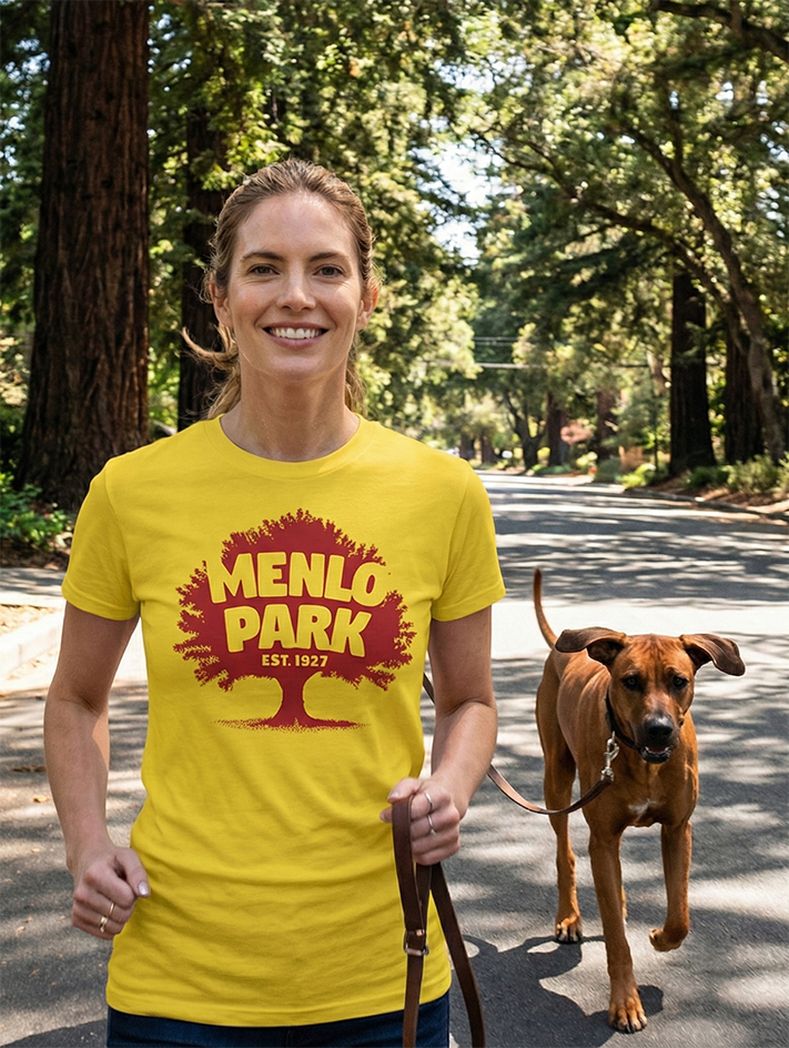 Woman wearing a yellow t-shirt with 'Menlo Park' design, standing with a dog on a leash on a tree lined street