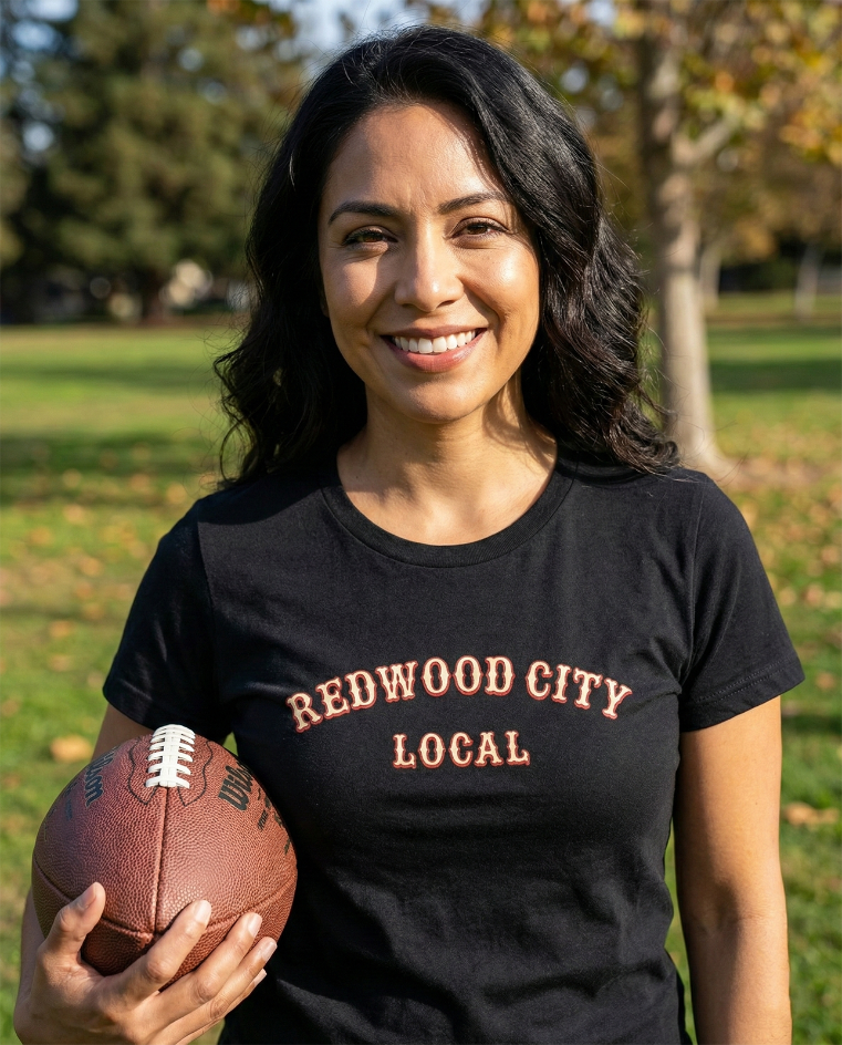 Woman wearing a Redwood City Local t-shirt holding a football and smiling in a park