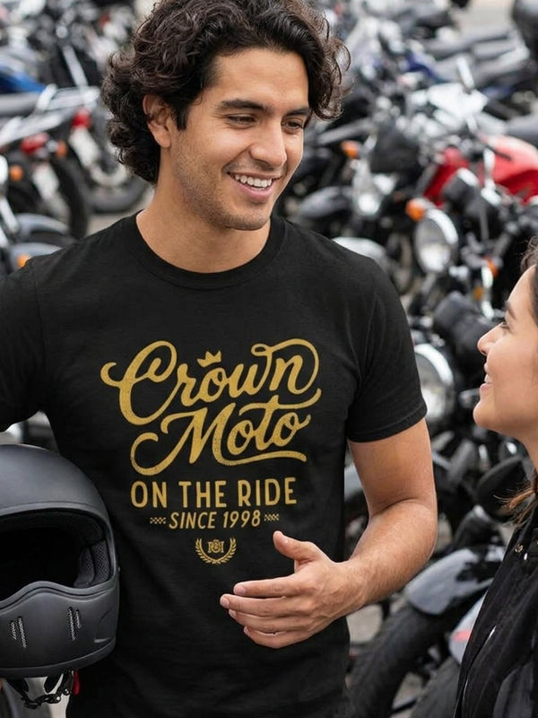 A man wearing a black t-shirt with a gold graphic that reads 'Crown Moto ON THE RIDE SINCE 1998' stands in a motorcycle shop with many bikes in the background.