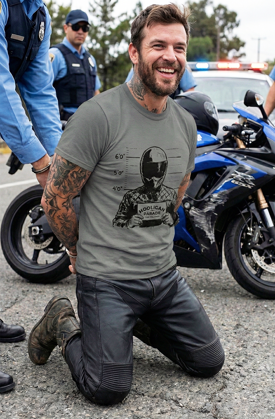 Man kneeling on the ground with tattoos, wearing a Hooligan Parade mug shot t-shirt, in front of motorcycles and police officers.