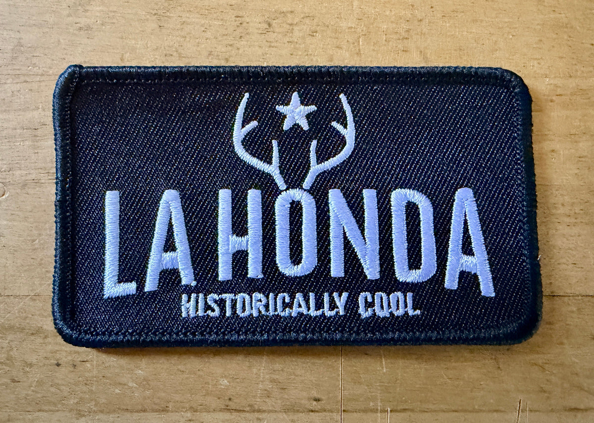 A rectangular embroidered patch with a black background and white text reading 'LA HONDA' and 'HISTORICALLY COOL,' plus a white antler-and-star graphic above, shown on a light wood surface.