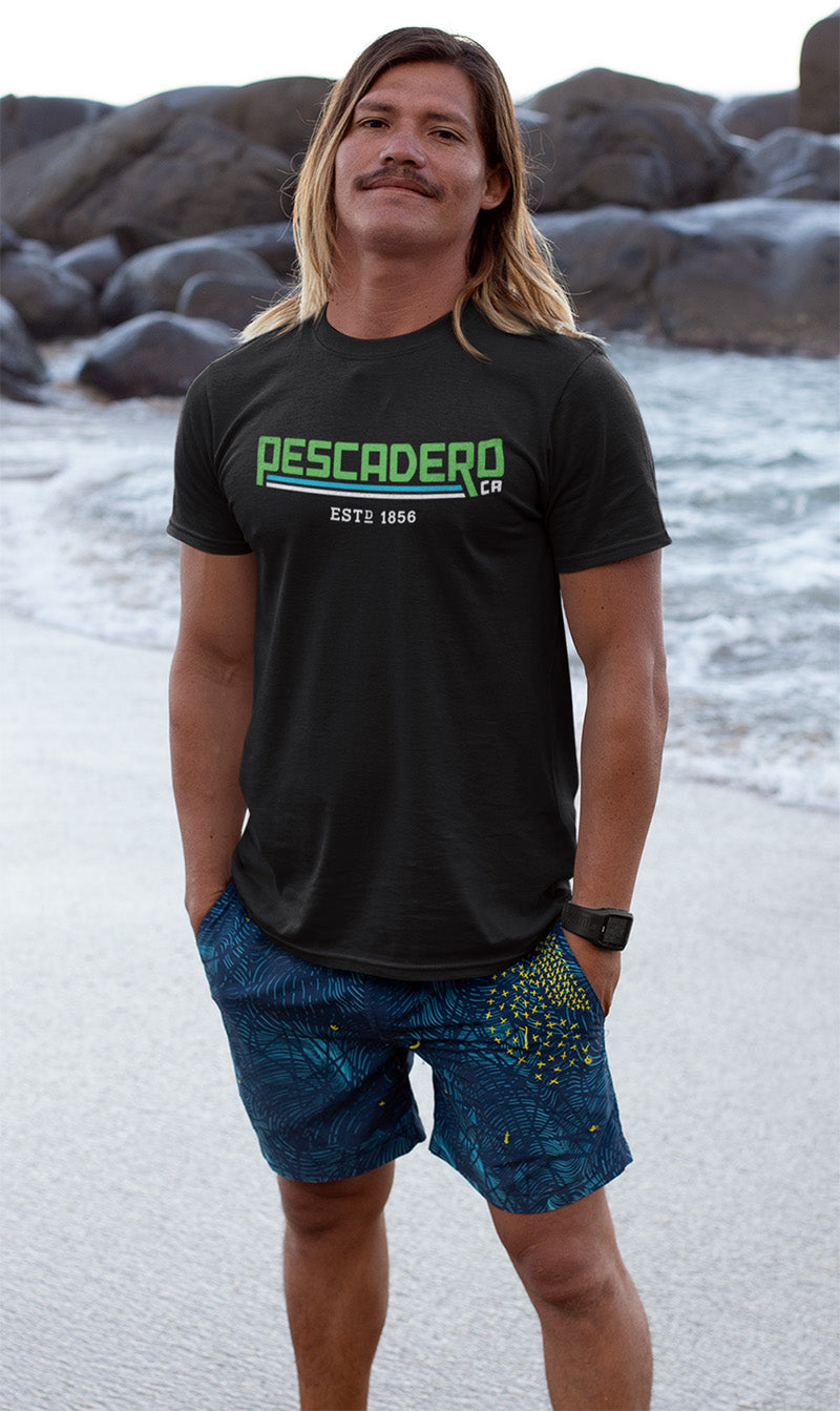 Man standing at the beach wearing a short sleeve t-shirt with original Pescadero design printed across the chest