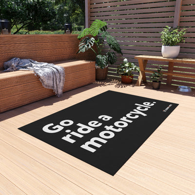 A rectangular rug with the phrase 'Go Ride a Motorcycle' printed in large white letters. The rug is placed on a wooden deck with outdoor furniture and a motorcycle in the background.