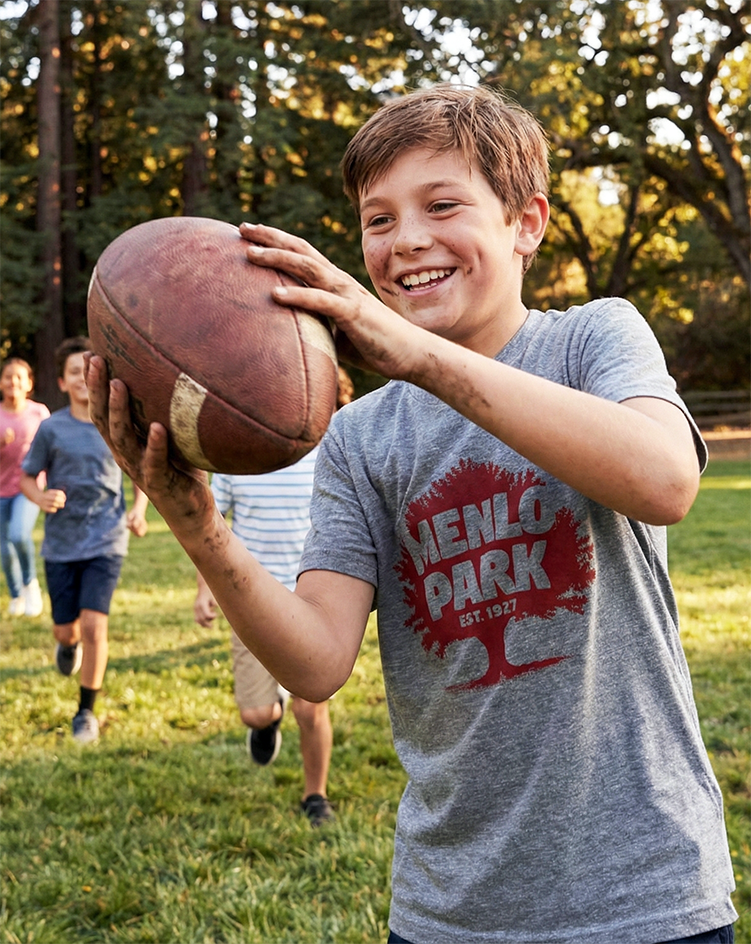 Boy wearing a Menlo Park t-shirt holding a football outdoors with friends in the background
