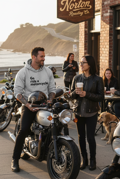 Man wearing a "Go ride a motorcycle" checkered flag hoodie sitting on a motorcycle with a woman holding a coffee cup in front of a coffee shop on the coast