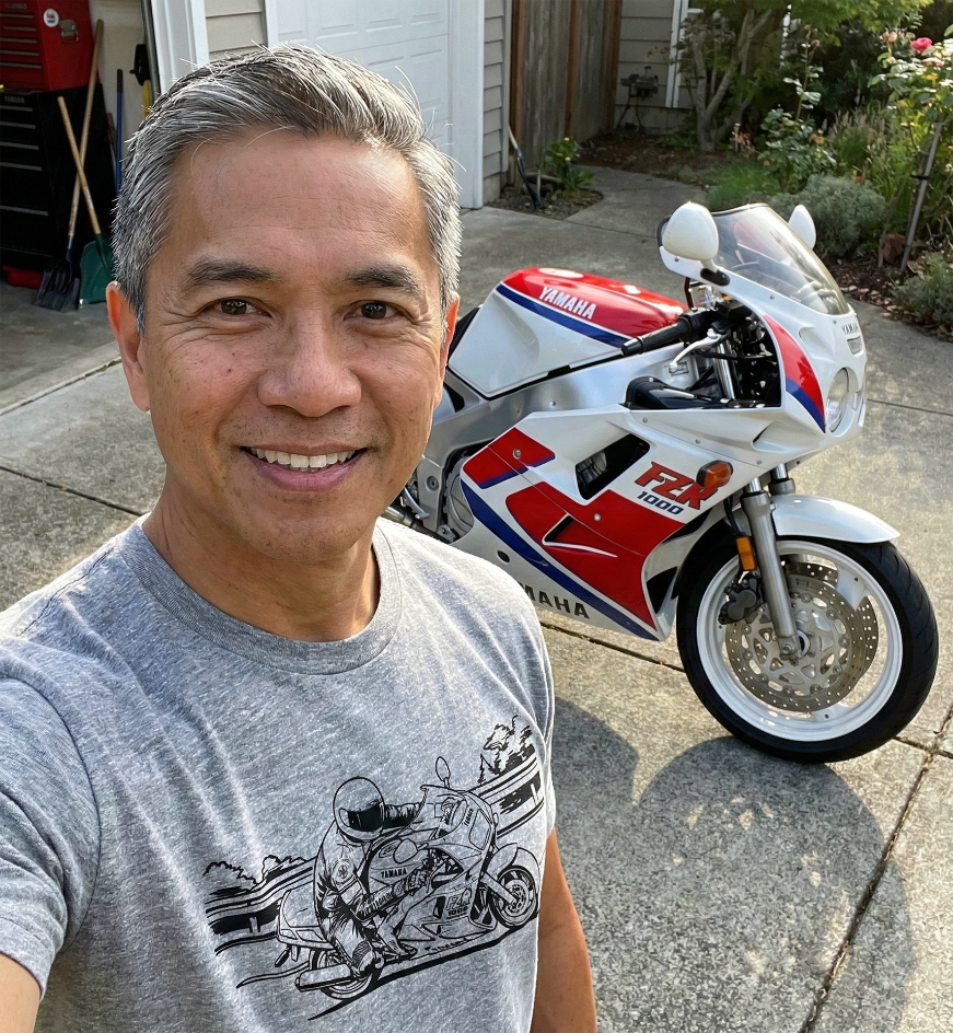Man taking a selfie with a red, white, and blue Yamaha FZR 1000 motorcycle in the background.