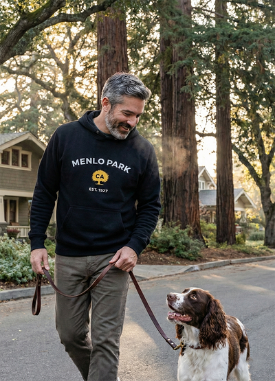 Man walking a dog on a leash in a Menlo Park neighborhood with redwood trees and houses in the background.