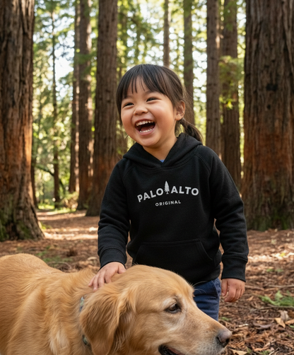 Child wearing a black hoodie with 'Palo Alto Original' text, standing next to a dog in a redwood forest.