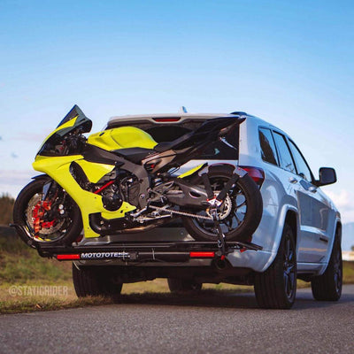 Yellow motorcycle on a trailer attached to a white SUV with a clear blue sky in the background.