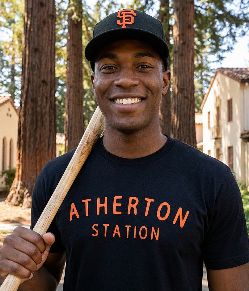 Man wearing a black t-shirt with orange 'Atherton Station' text and a SF Giants baseball cap, holding a baseball bat, standing in a forested area.