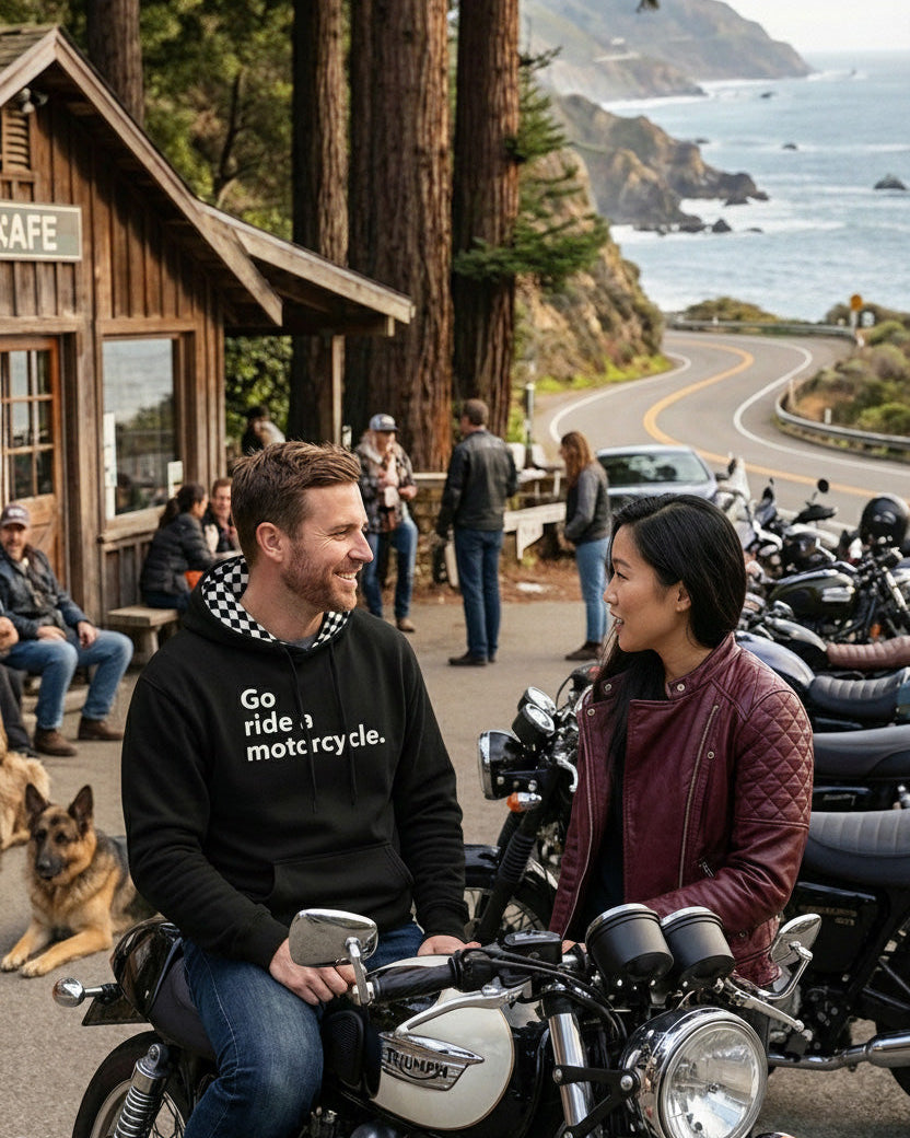 Man wearing a "Go ride a motorcycle" Checkered Flag Hoodie talking to a woman in front of a cafe on the California coast with motorcycles, people and dogs in the background