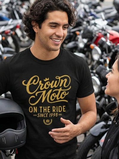 A man wearing a black t-shirt with a gold graphic that reads 'Crown Moto ON THE RIDE SINCE 1998' stands in a motorcycle shop with many bikes in the background.