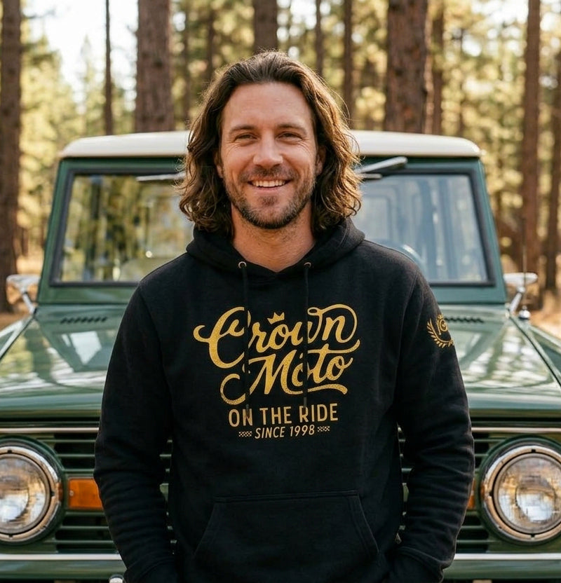 A smiling man with shoulder-length brown hair stands in front of a green vintage vehicle in a forest, wearing a black hoodie with a gold 'Crown Moto' graphic on the chest.