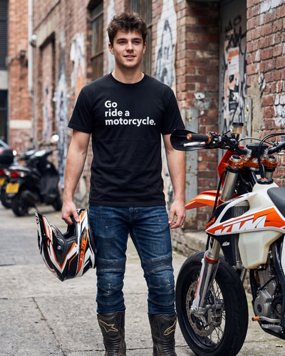 A man wearing a black graphic t-shirt with the white text 'Go ride a motorcycle.' stands in an urban alleyway next to an orange and white KTM motorcycle, holding a helmet.