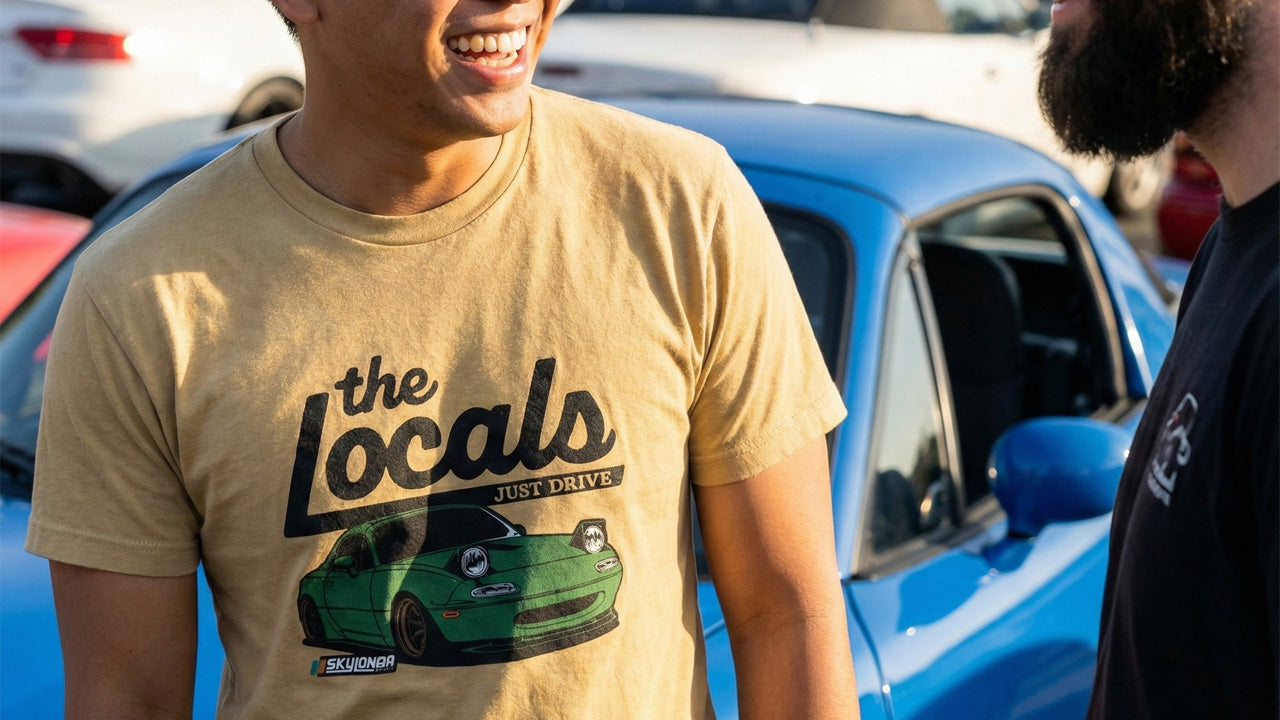 Person wearing a 'the Locals' t-shirt with a Miata graphic, standing next to a blue car.