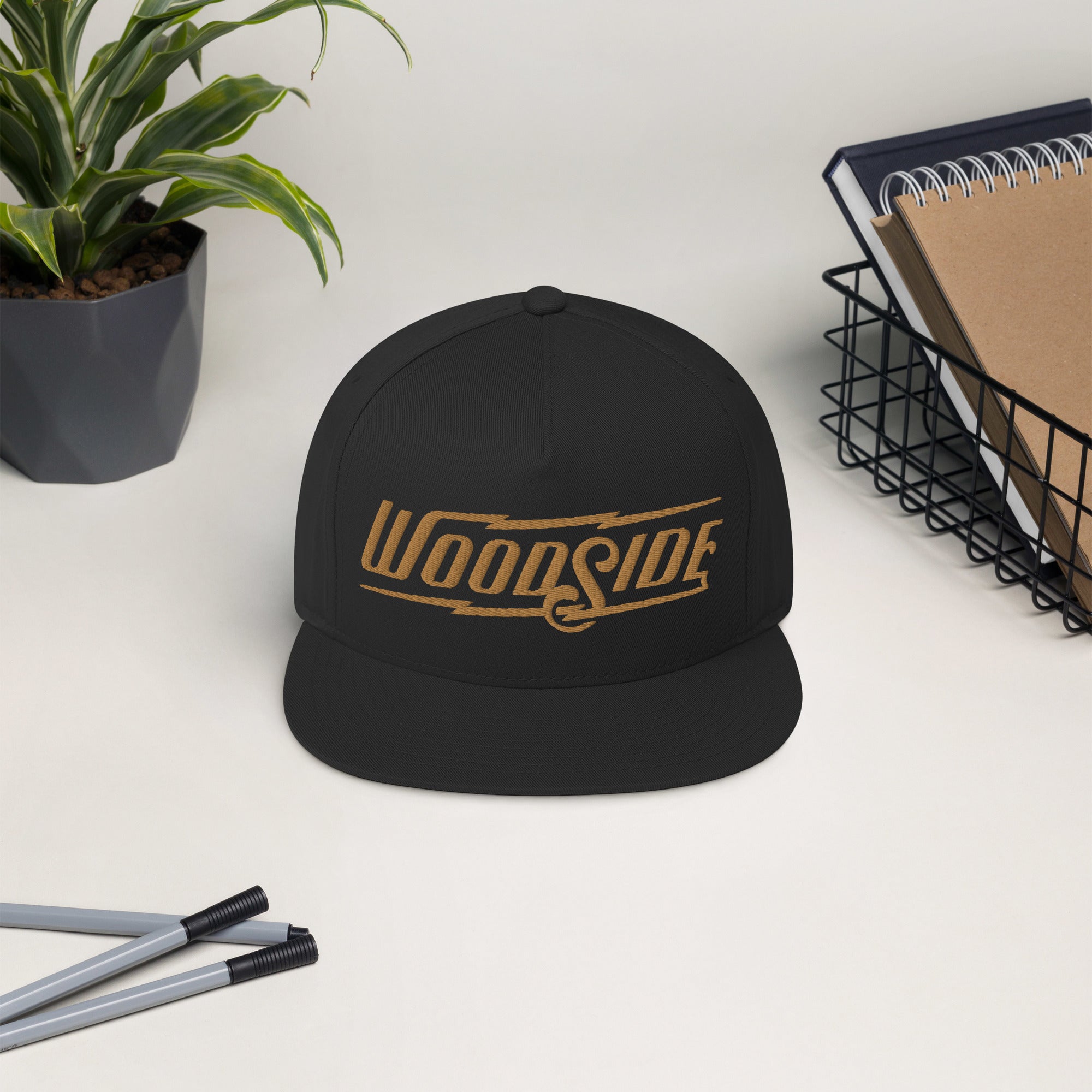 Black Woodside Brand embroidered snapback hat with gold embroidery displayed on a desk