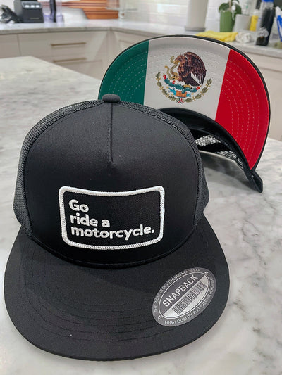 Black cap with 'Go ride a motorcycle.' text on a marble surface with a Mexican flag printed on underside of the brim