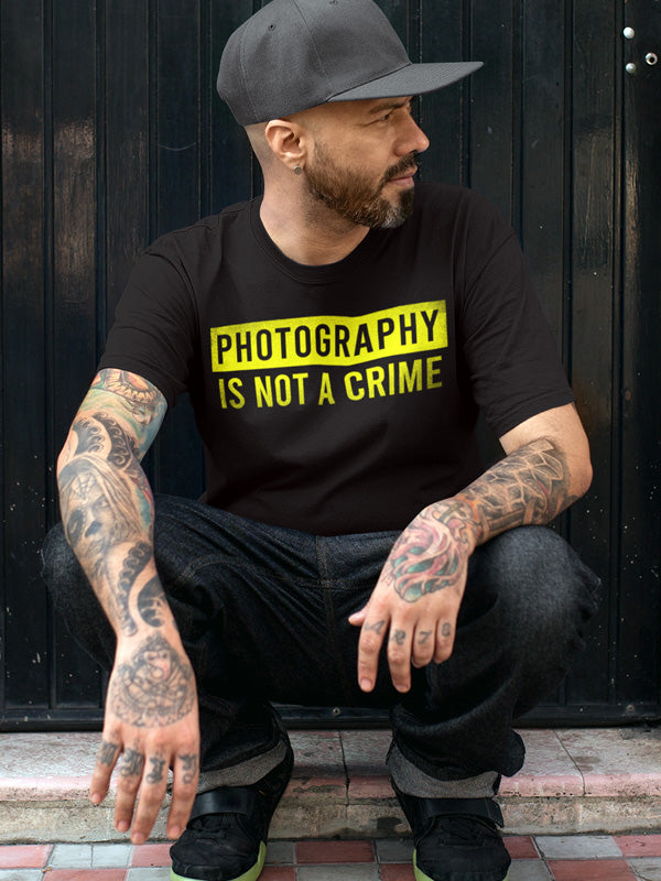 Tattooed man sitting wearing hat and short sleeve t-shirt with "photography is not a crime" in yellow on chest