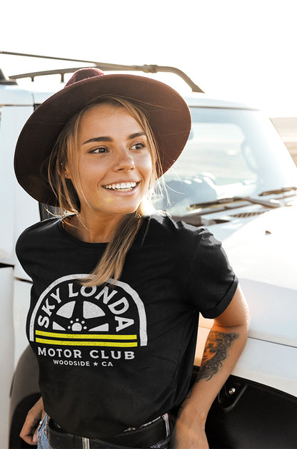 Young woman leaning against a jeep wearing a black t-shirt with "Sky Londa Motor Club" wheel logo on chest