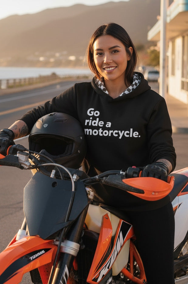 Woman wearing a hoodie with 'Go ride a motorcycle' text, sitting on a motorcycle by a scenic road.