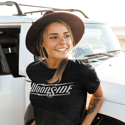 Young woman smiling leaning against jeep wearing a black t-shirt with original Woodside logo printed across chest