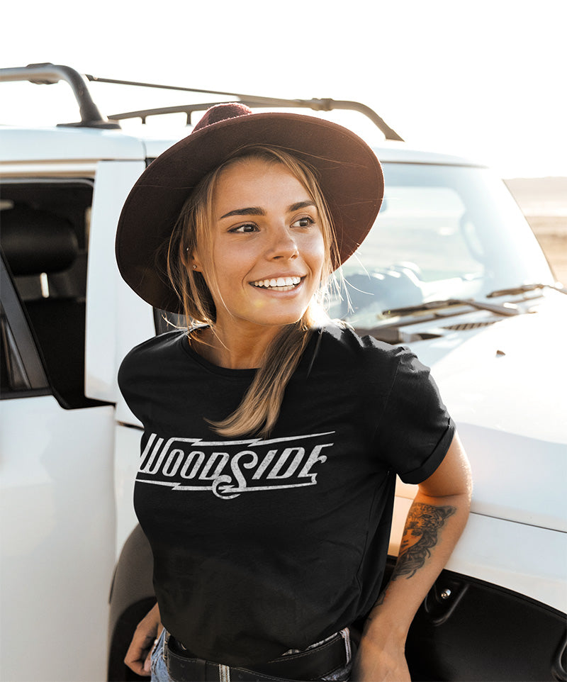 Young woman smiling leaning against jeep wearing a black t-shirt with original Woodside logo printed across chest